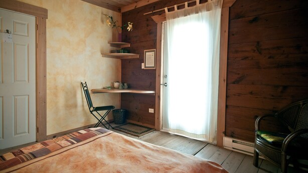 A wooden room with a bed, a chair, a shelf with a vase, and a door.