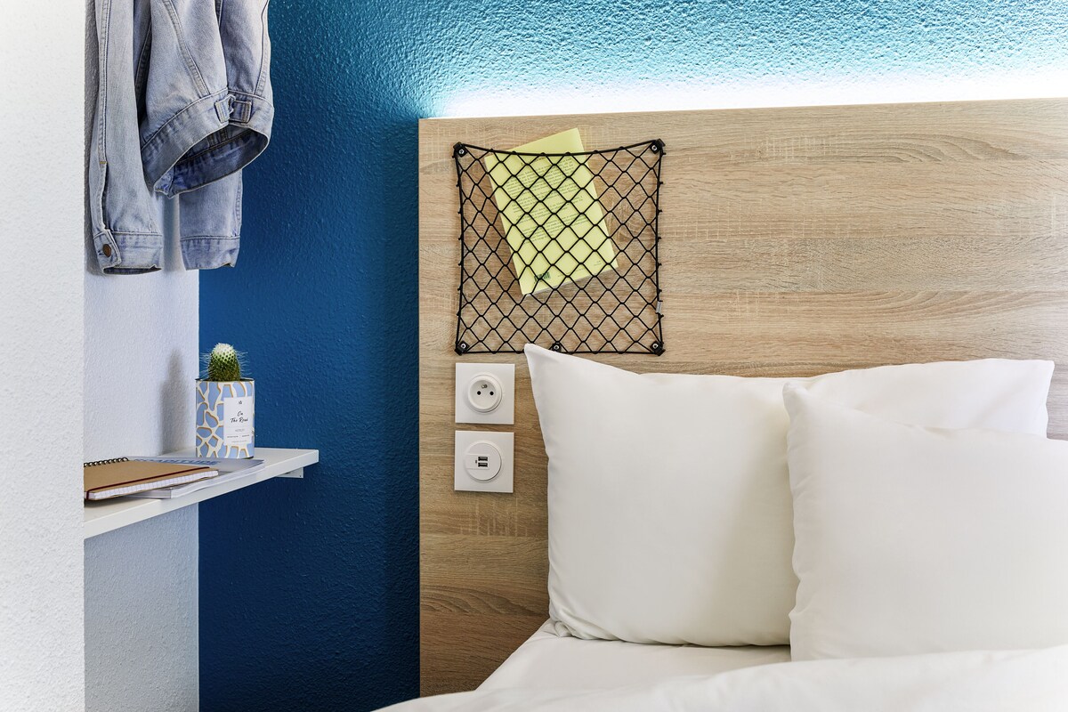 A neatly arranged hotel room with a wooden headboard, a white bed, a shelf with a notebook, a small cactus, and a denim jacket hanging on the wall.