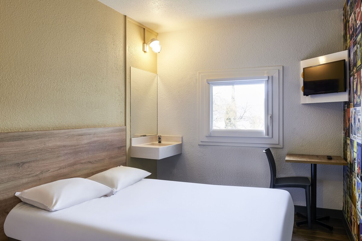 A hotel room with a bed, a bedside table, a mirror, a sink, a chair, and a TV mounted on the wall.