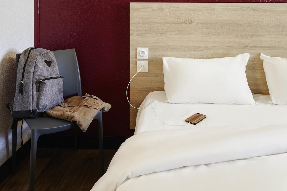 A hotel room with a bed, a chair, a backpack, and a wooden headboard.