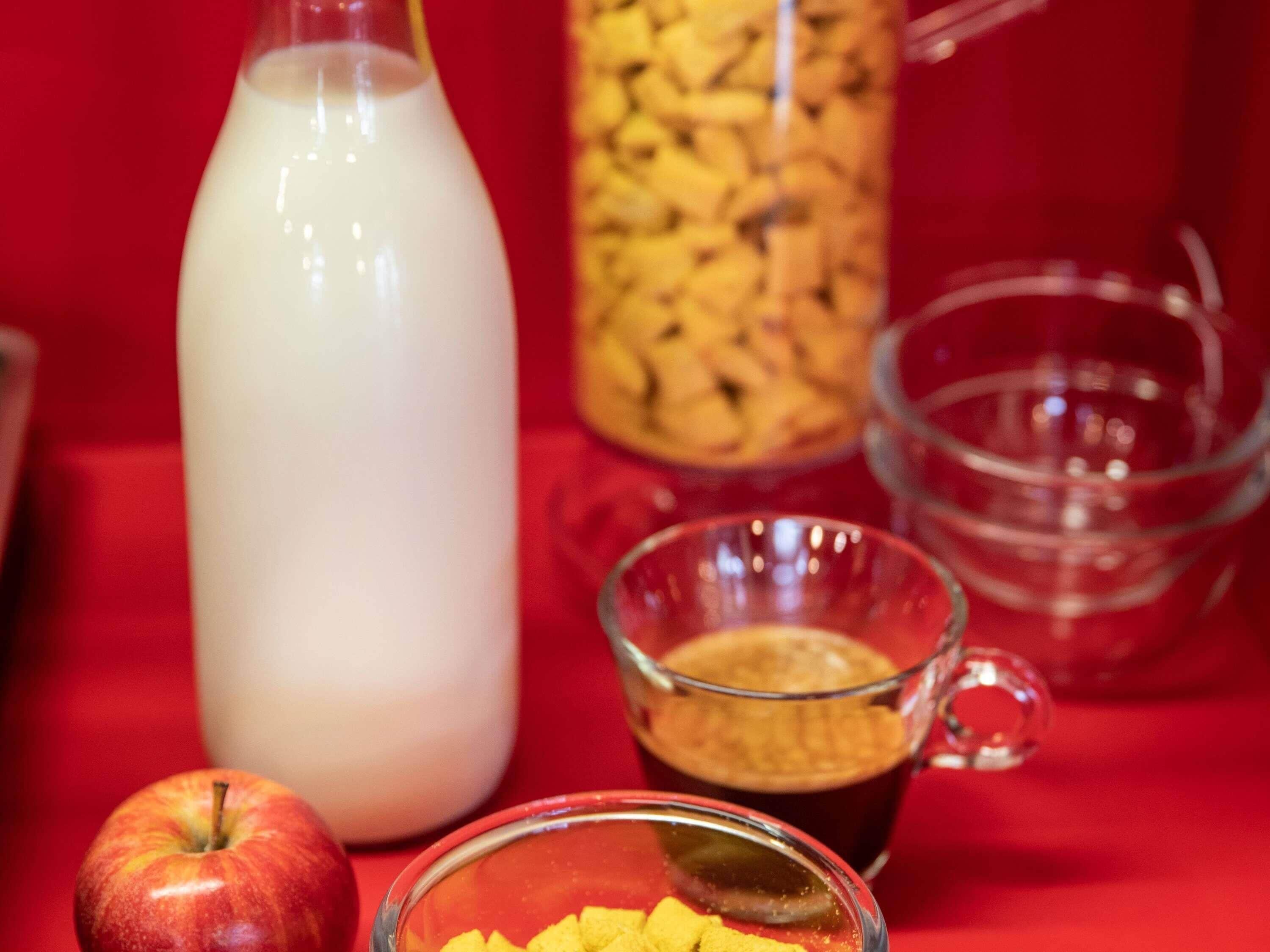 Een fles melk, een mok met donkere vloeistof, een kom gele blokjes en een appel op een rood oppervlak.