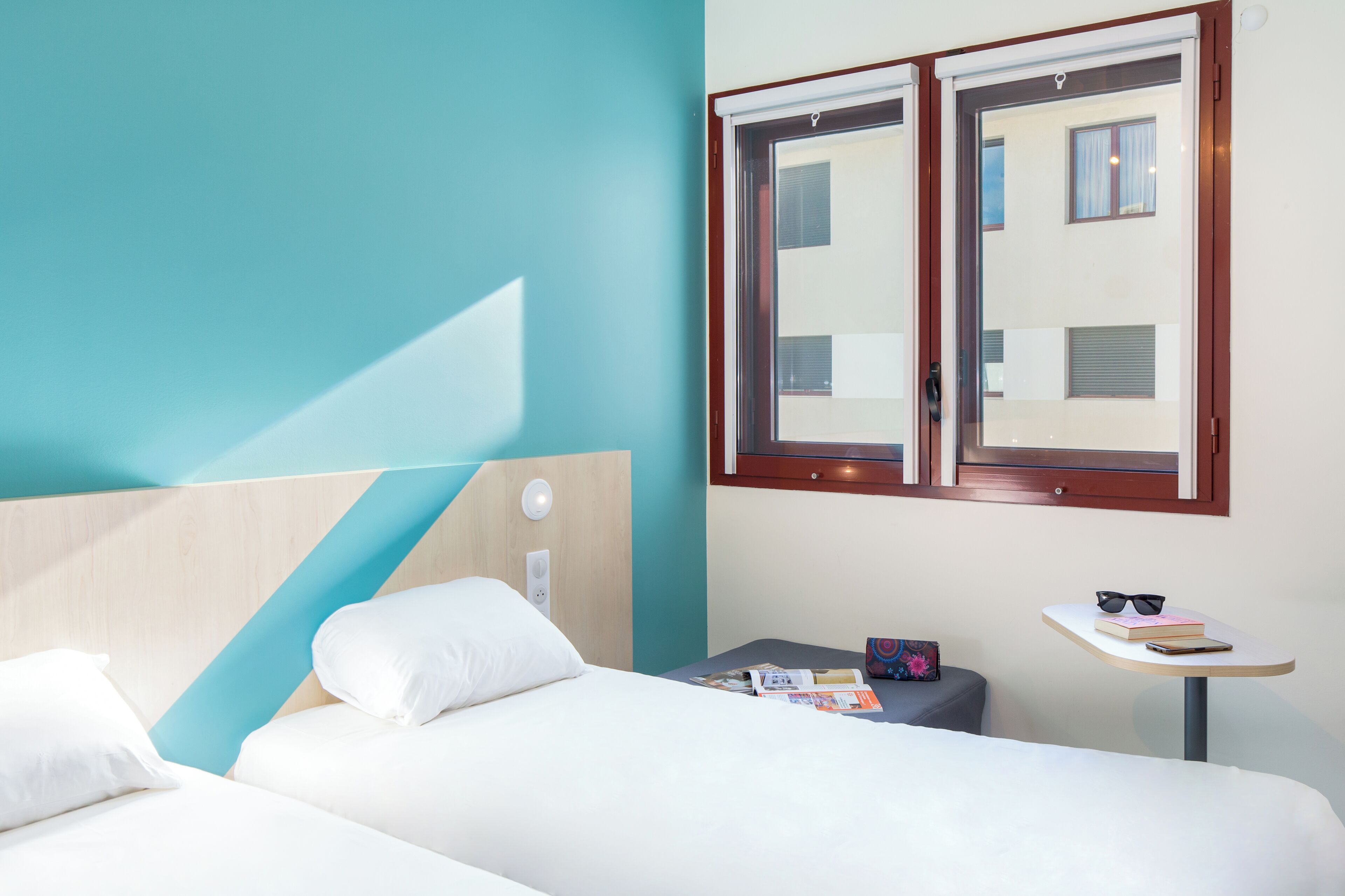 A hotel room with a bed, a small table with a book and sunglasses, and a window with a view of buildings.