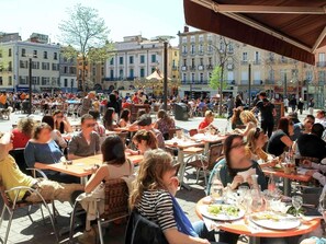 Un café al aire libre con mesas y sillas, rodeado de edificios con señalización.