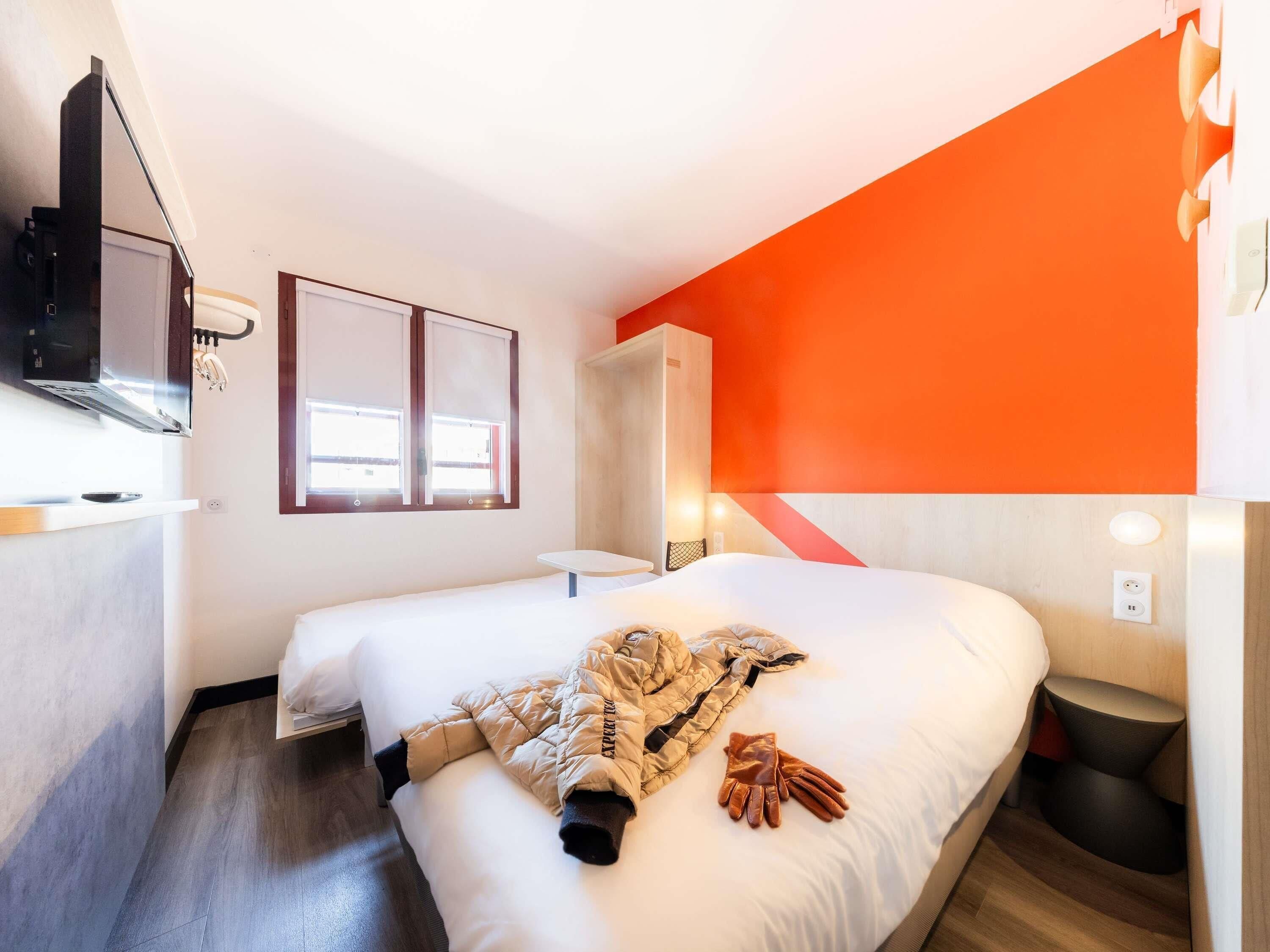 A hotel room with a bed, a TV, a window, and a wall with an orange accent.
