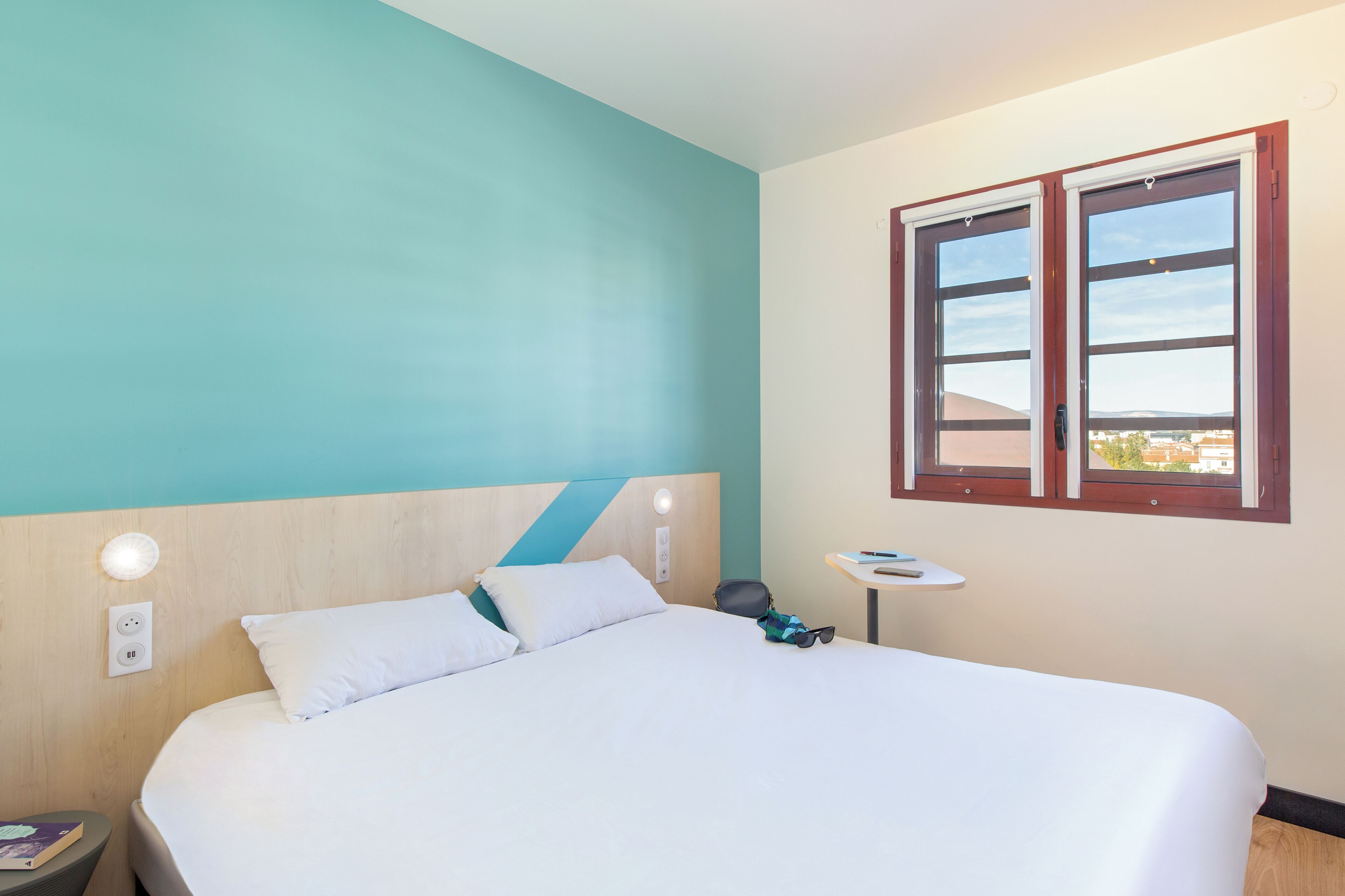 Una habitación de hotel con cama, mesita de noche, una pequeña mesa y ventana con vistas.