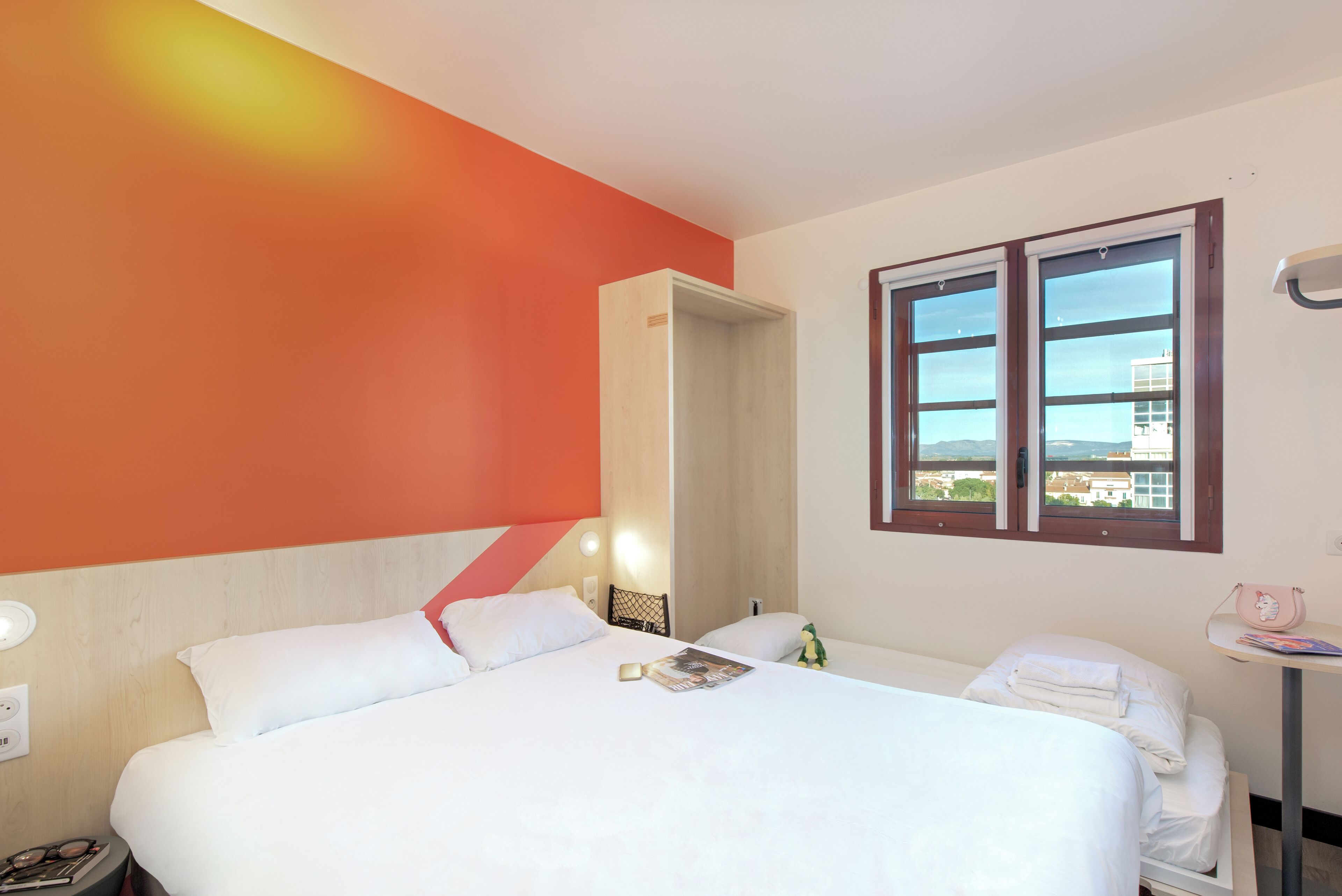 A hotel room with a bed, a nightstand, a table with a lamp, and a window with a view of buildings.