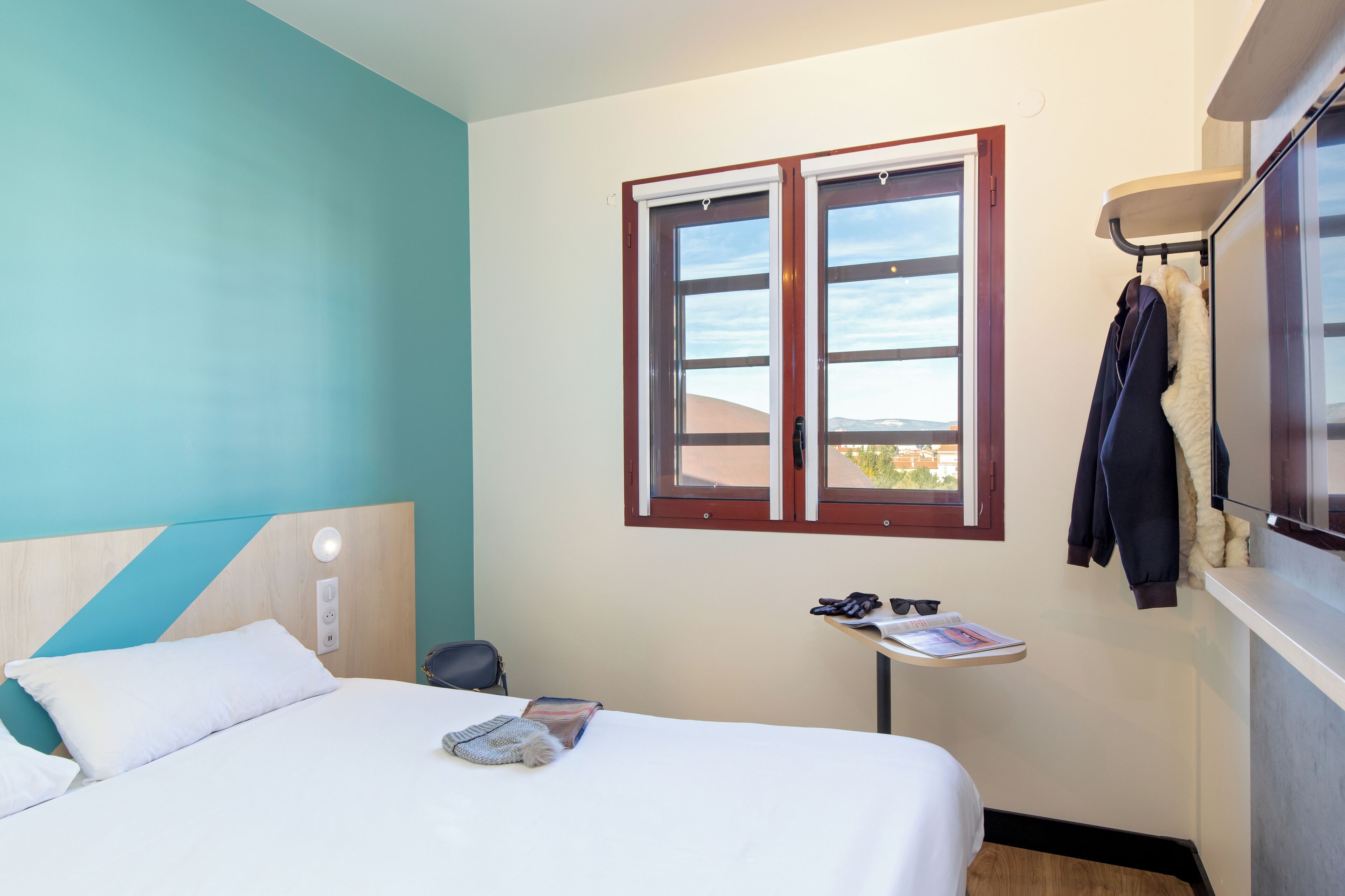 A hotel room with a bed, a small table with a magazine, a coat rack with a jacket, and a window with a view of the outside.