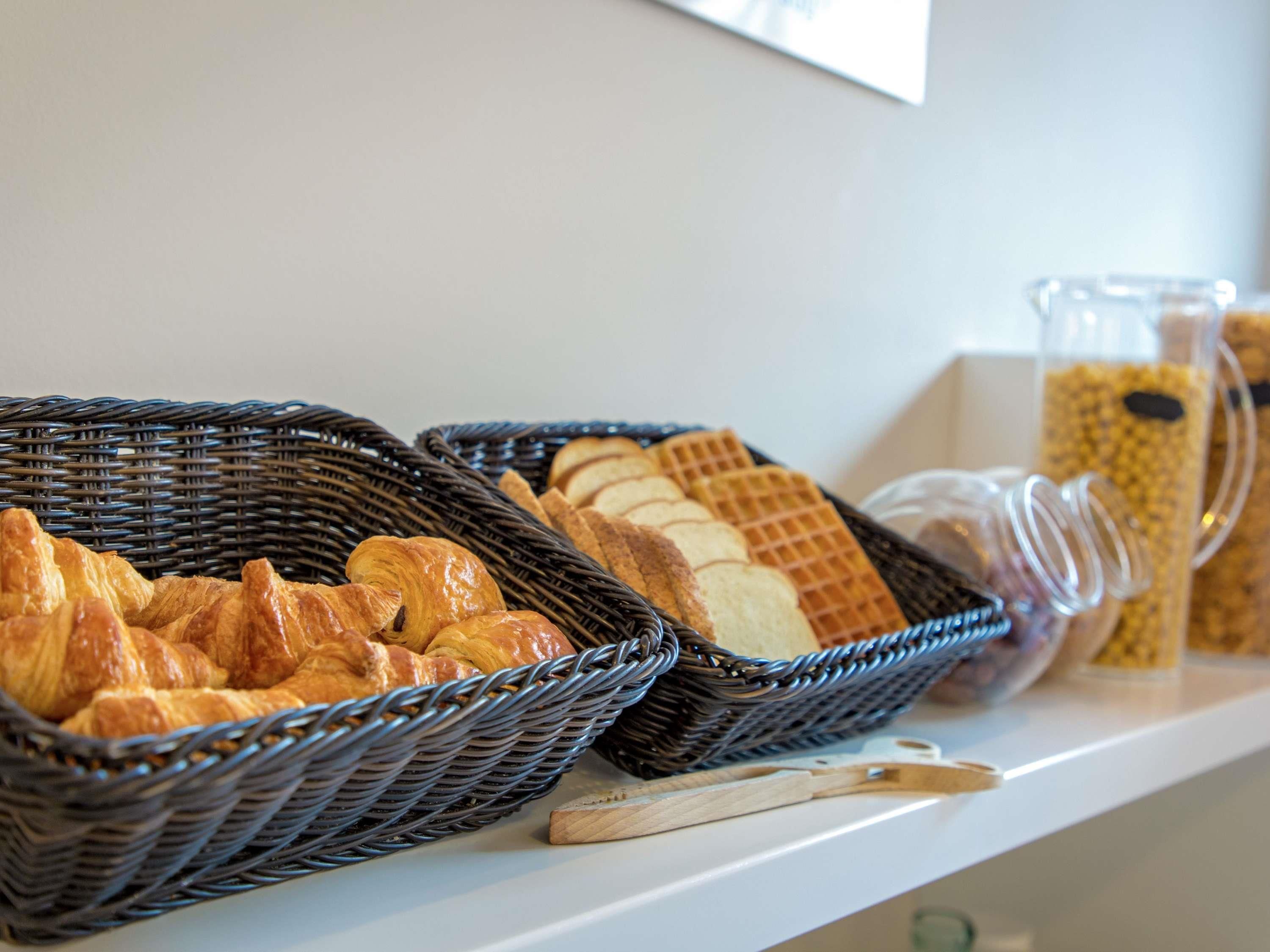 A breakfast buffet with baskets of croissants and waffles, a glass pitcher, and a wooden cutting board.