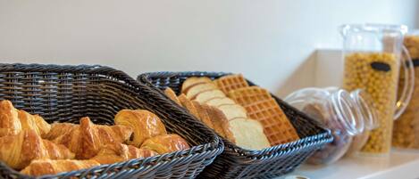 A breakfast buffet with baskets of croissants and waffles, a glass pitcher, and a wooden cutting board.