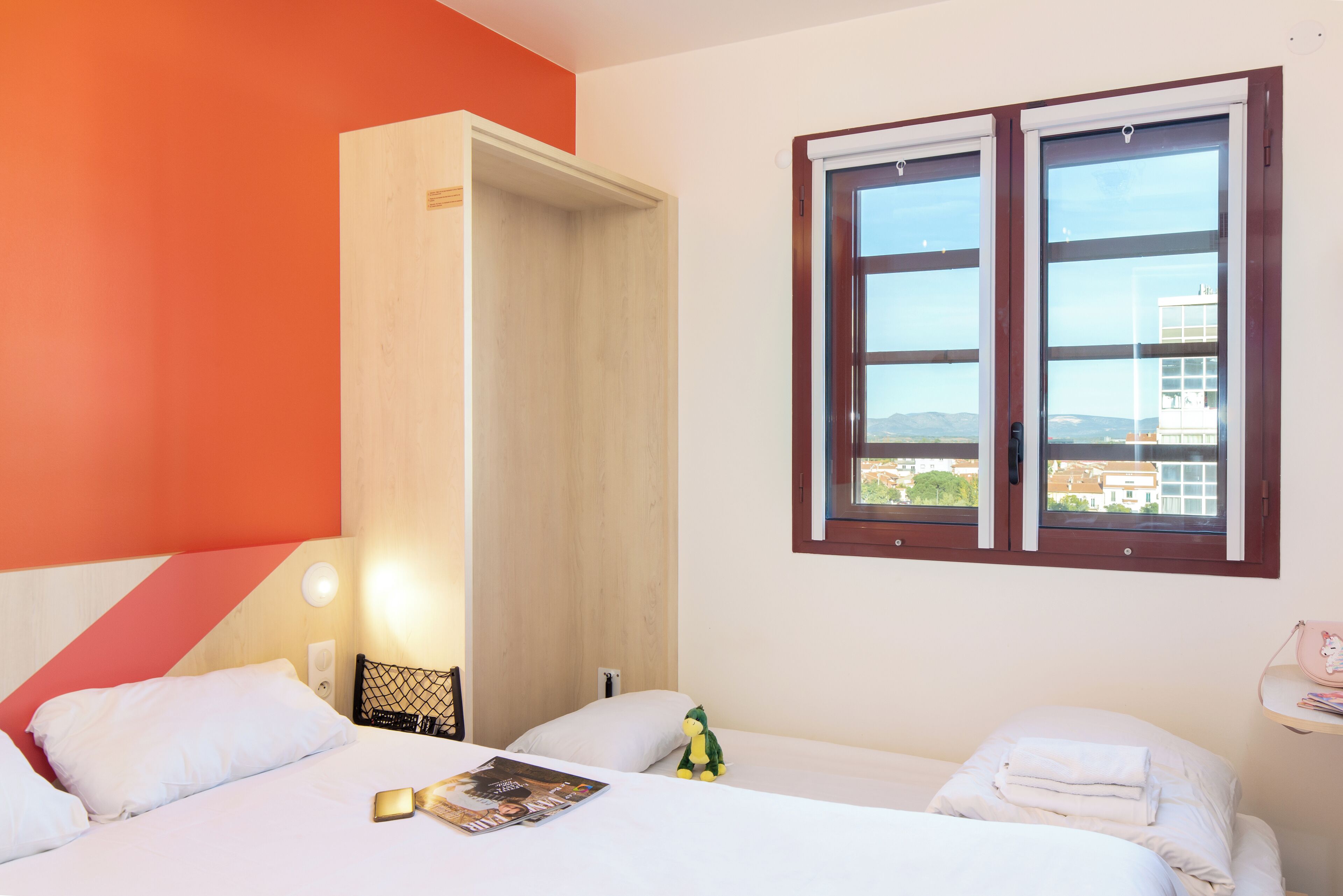 Habitación de hotel con dos camas, un armario de madera, una ventana con vistas a edificios y una pared con diseño geométrico.