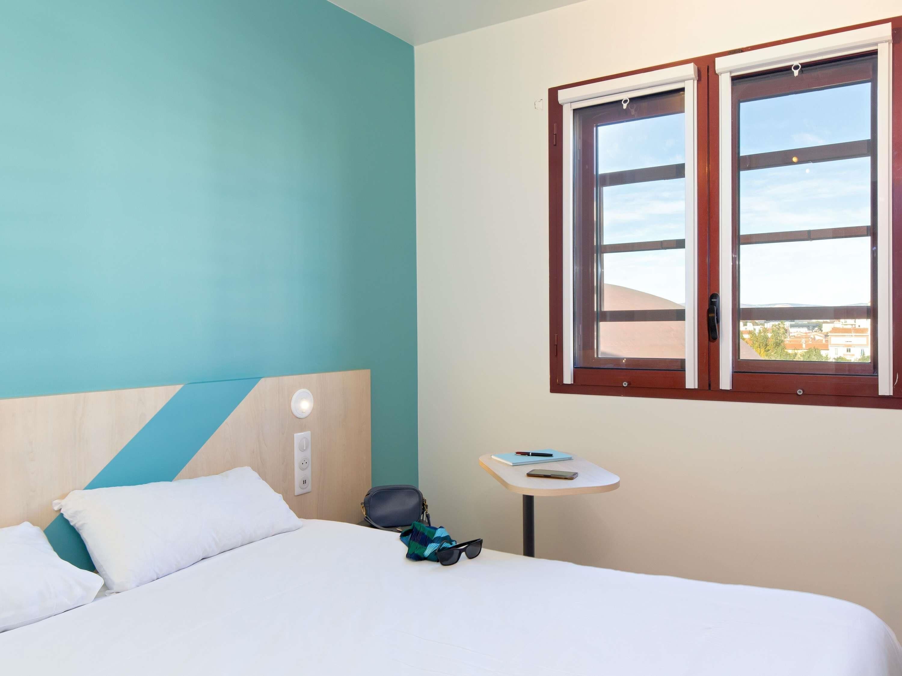 Habitación de hotel con cama, una mesita, ventana con vistas y pared con diseño geométrico.