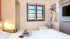 Habitación de hotel con cama, ventana con persianas, televisor en la pared, mesita con lámpara y perchero con chaqueta.