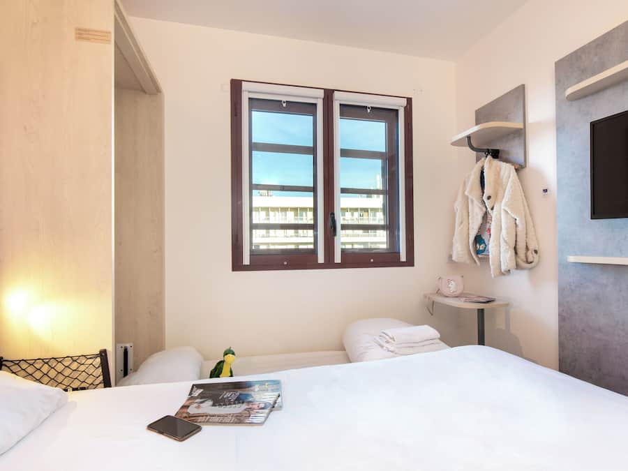 A hotel room with a bed, a window with blinds, a TV mounted on the wall, a small table with a lamp, and a coat rack with a jacket.