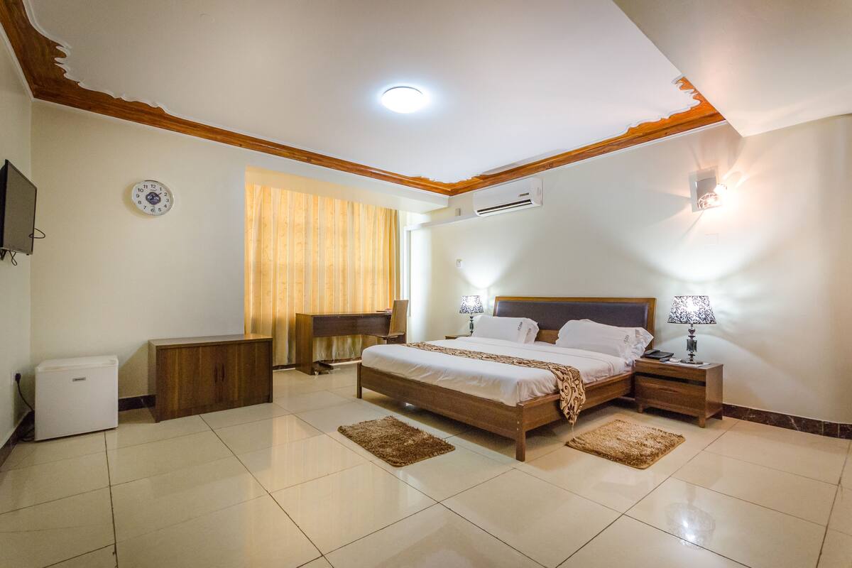 A bedroom with a bed, bedside tables, a desk, a chair, a TV, and an air conditioning unit.