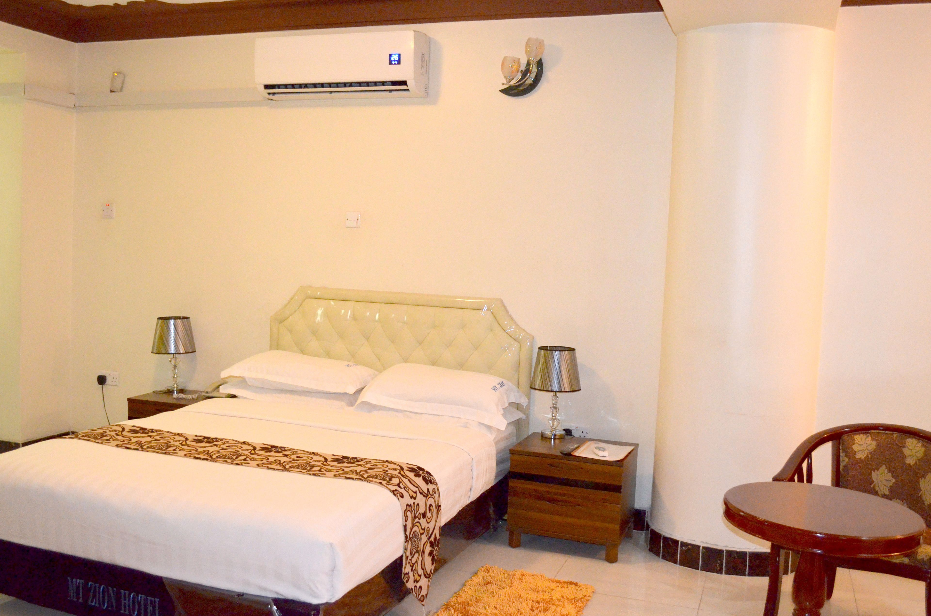 A hotel room with a bed, bedside tables, a chair, an air conditioning unit, and a lamp.