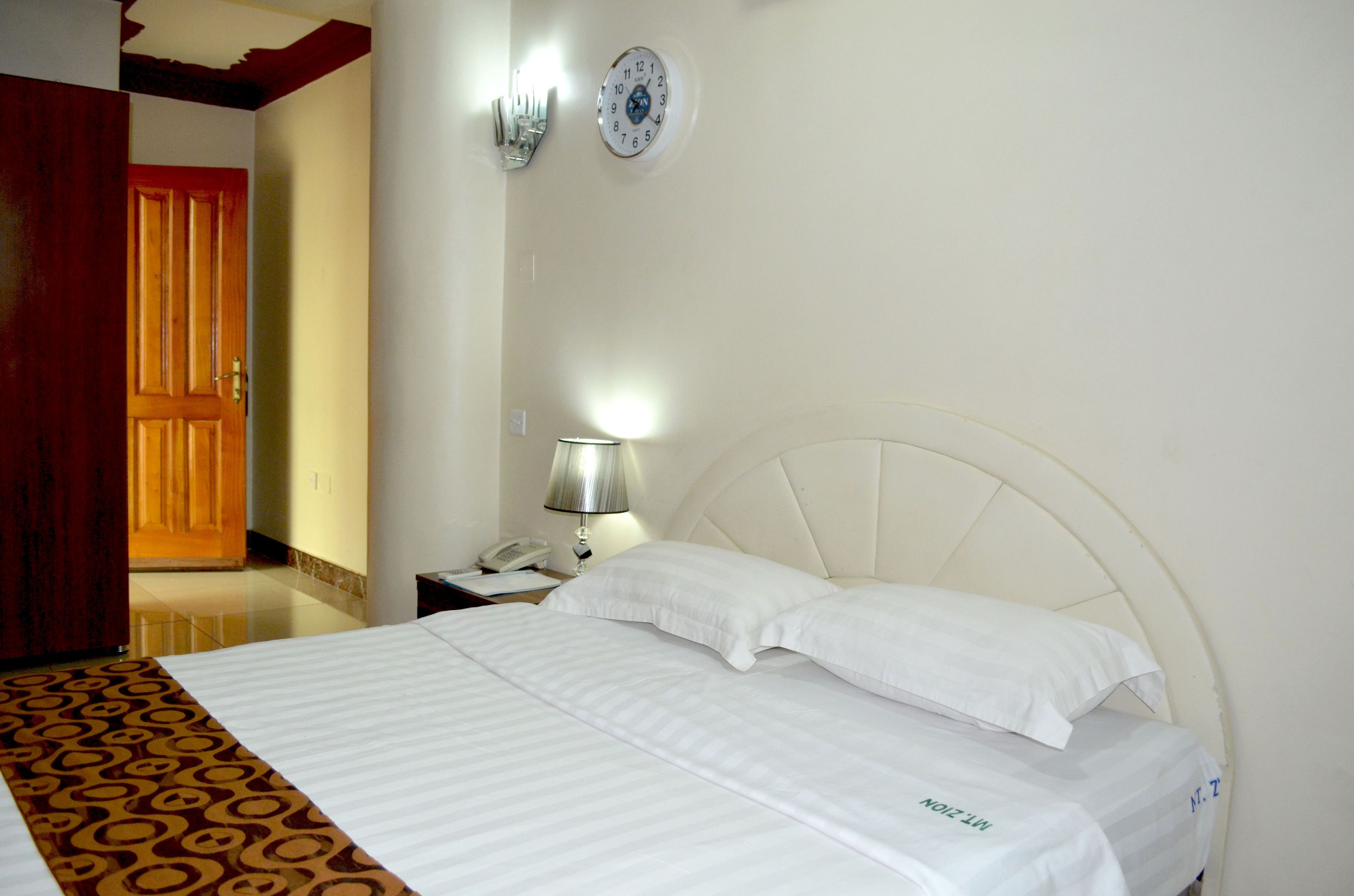A hotel room with a bed, a nightstand with a lamp, a wall clock, and a door.