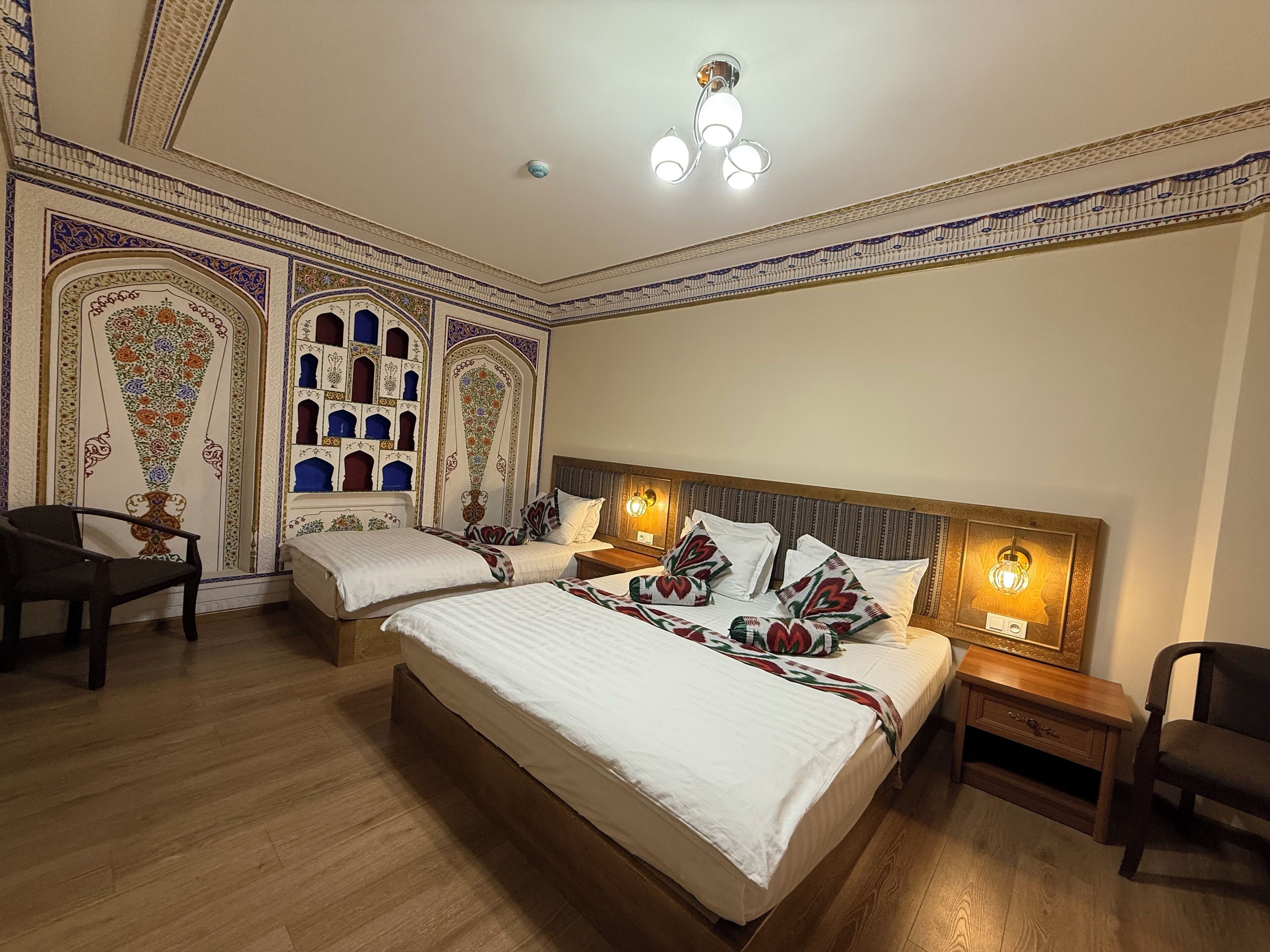 A hotel room with two beds, a wooden floor, decorative wall panels, and a ceiling light fixture.