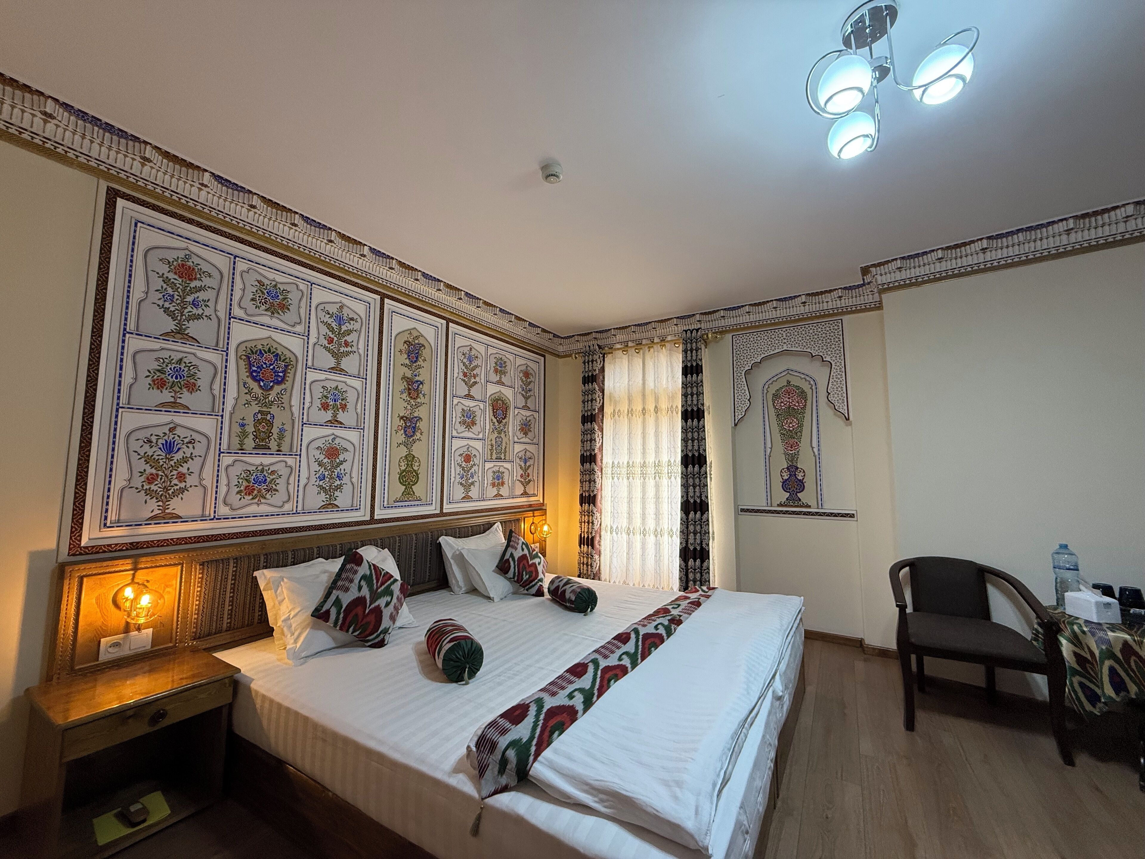 A hotel room with a large bed, a nightstand, a chair, and a wall decorated with intricate patterns.