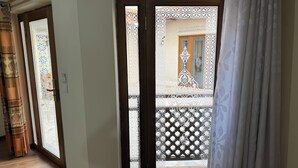 A room with a wooden floor, a door with intricate white patterns, and a view of a balcony with a lattice design. - Hotel Fatima (Bukhara)