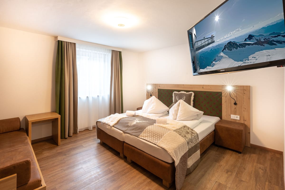 A hotel room with a wooden floor, a bed with a headboard, a bedside table, a TV mounted on the wall, and a window with curtains.