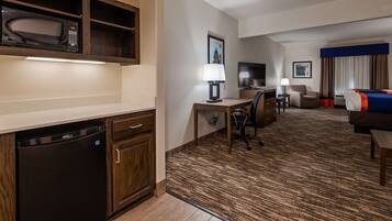A hotel room with a kitchenette, a desk with a chair, a television, a bed, and a carpeted floor.