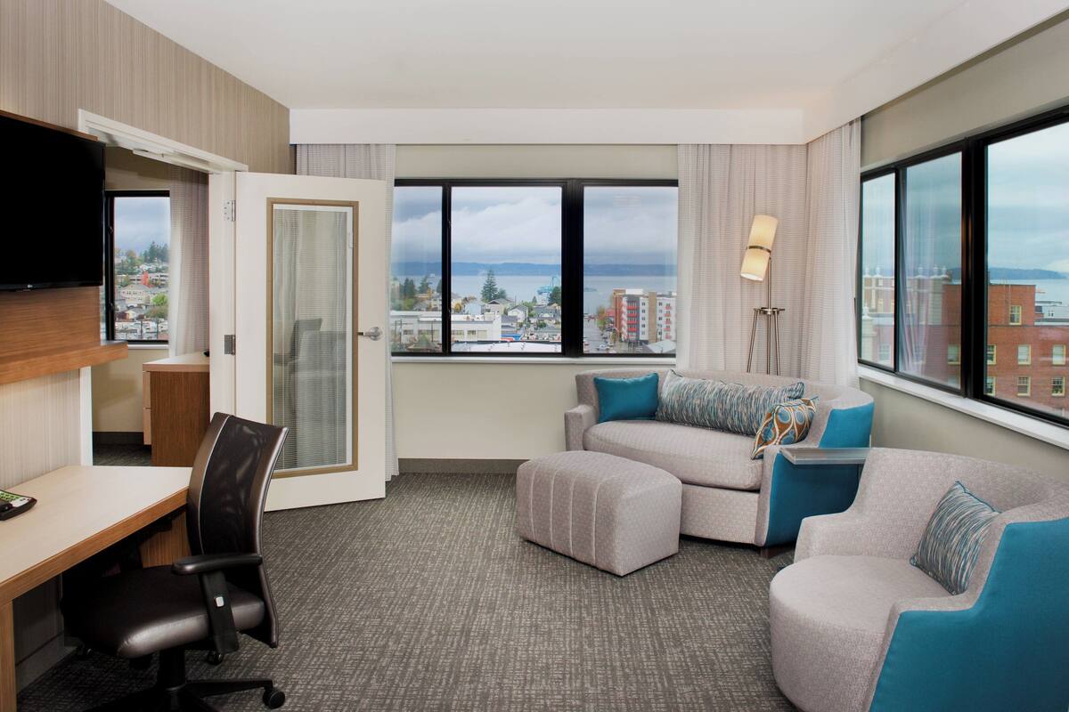 Ein Hotelzimmer mit Schreibtisch, Bürostuhl, zwei Sofas und einem großen Fenster mit Blick auf die Stadt.