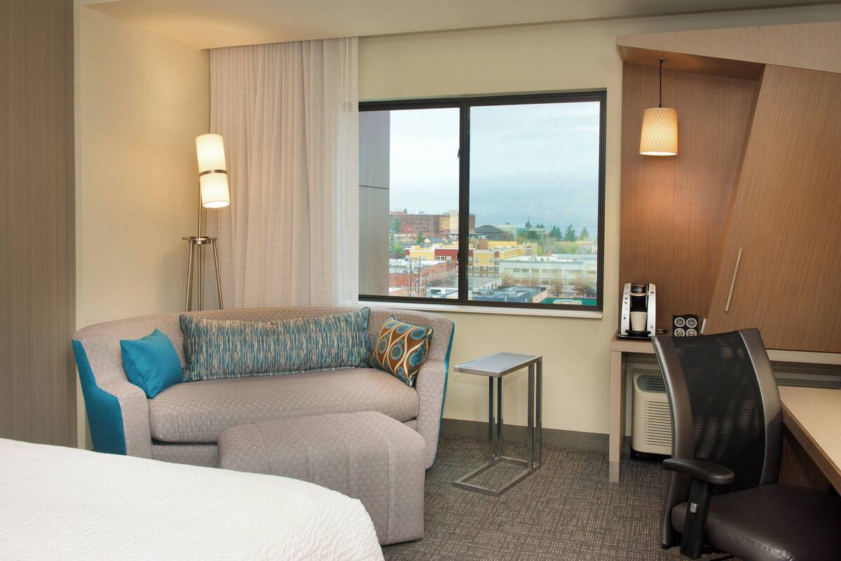Ein Hotelzimmer mit einer Sitzecke, einem kleinen Tisch, einem Sessel, einem Bett, einem Fenster mit Blick auf die Stadt und einer Lampe.