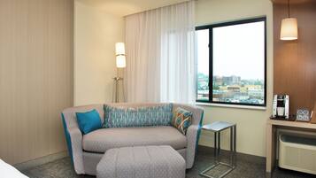 Habitación de hotel con un sofá gris, cojines estampados, una mesita auxiliar, una cafetera y una ventana con vistas a la ciudad.