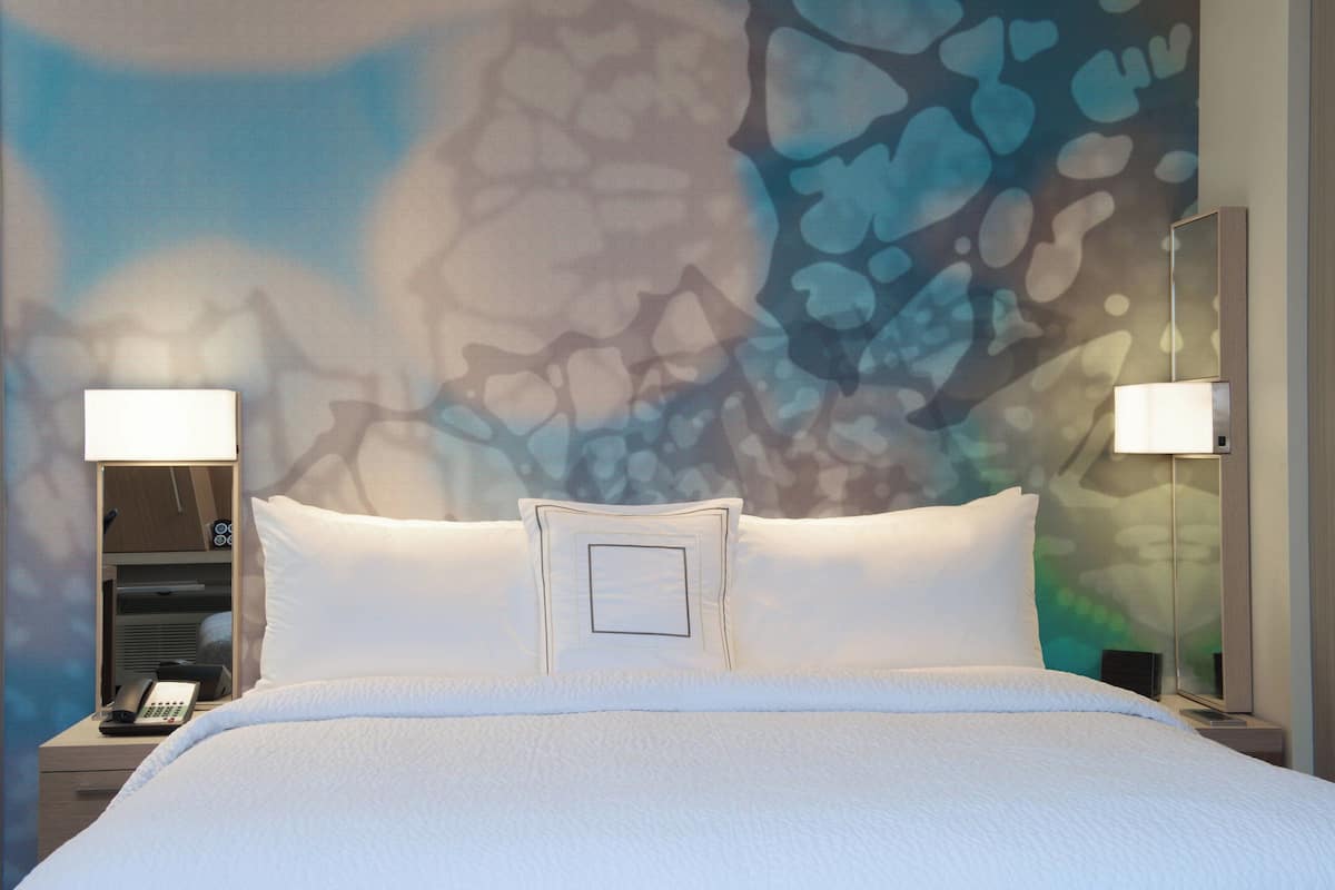 Ein Hotelzimmer mit einem großen Bett, zwei Nachttischlampen, einem Nachttisch mit Telefon und einer Wand mit strukturierter, abstrakter Gestaltung.
