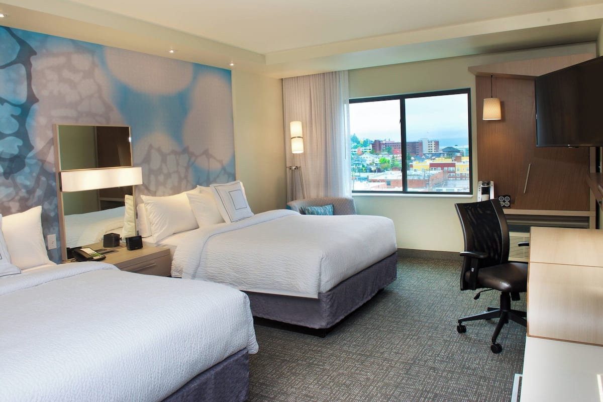 Ein Hotelzimmer mit zwei Betten, einem Schreibtisch, einem Stuhl, einem großen Fenster mit Stadtblick und einer Wand mit einer strukturierten Wandmalerei.
