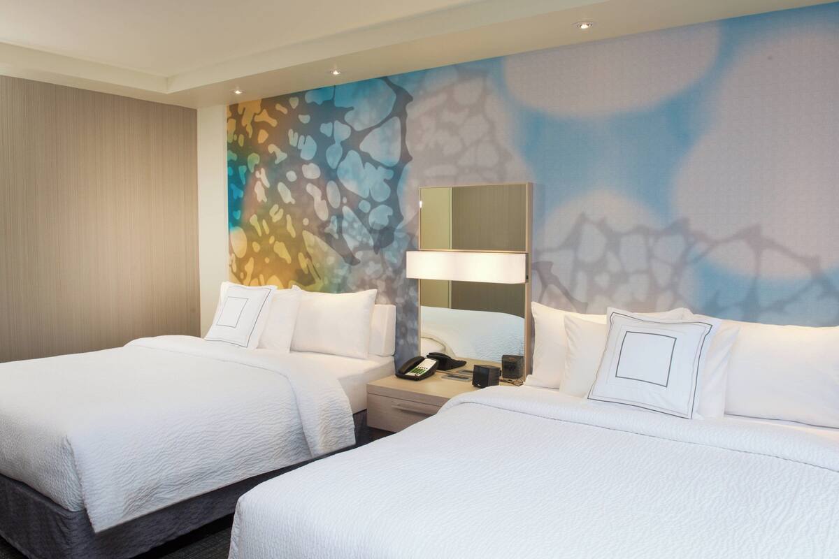 Ein Hotelzimmer mit zwei Betten, einem Nachttisch mit Lampe, Telefon und einer Wand mit einem farbenfrohen abstrakten Design.
