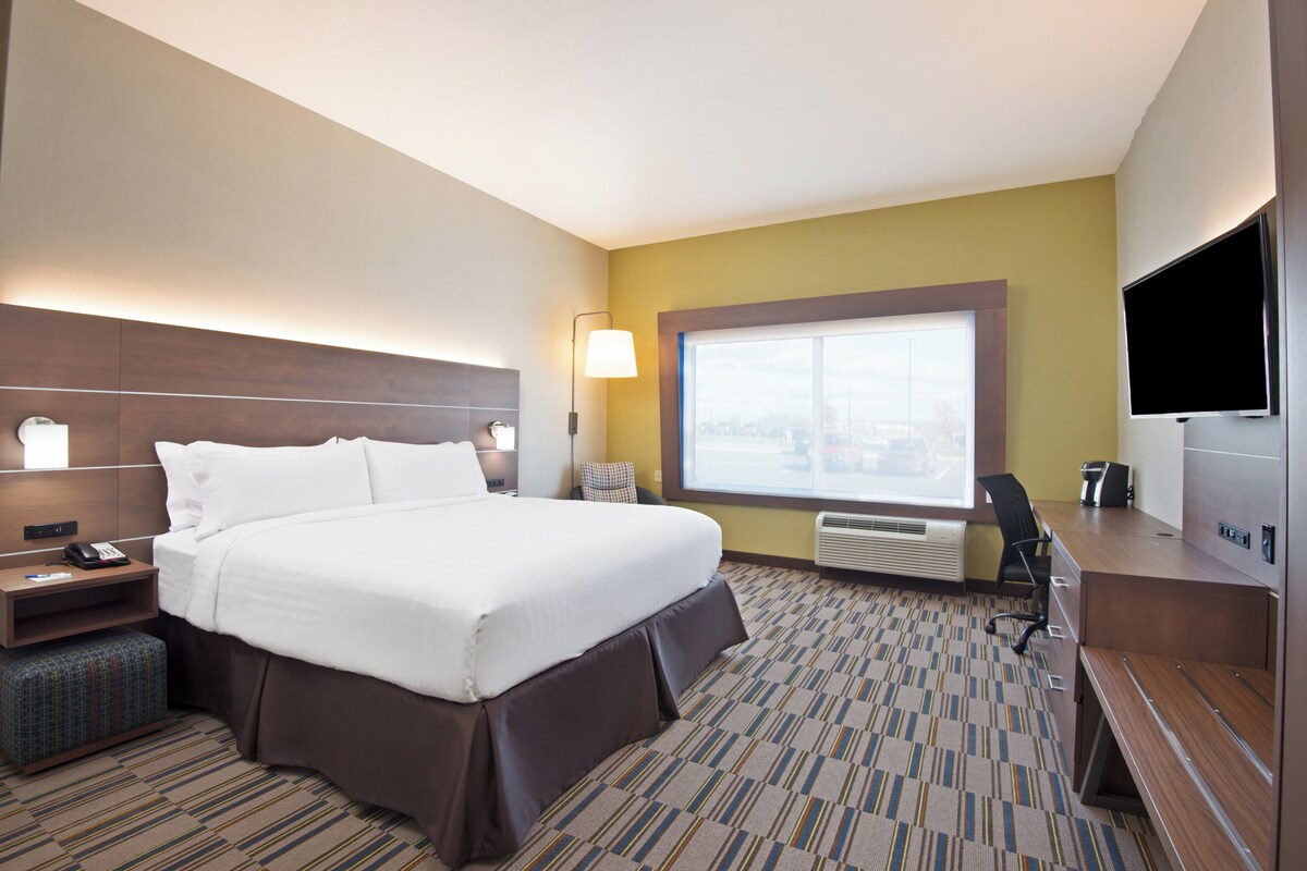 A hotel room with a large bed, a desk, a chair, a TV, and a window with a city view.