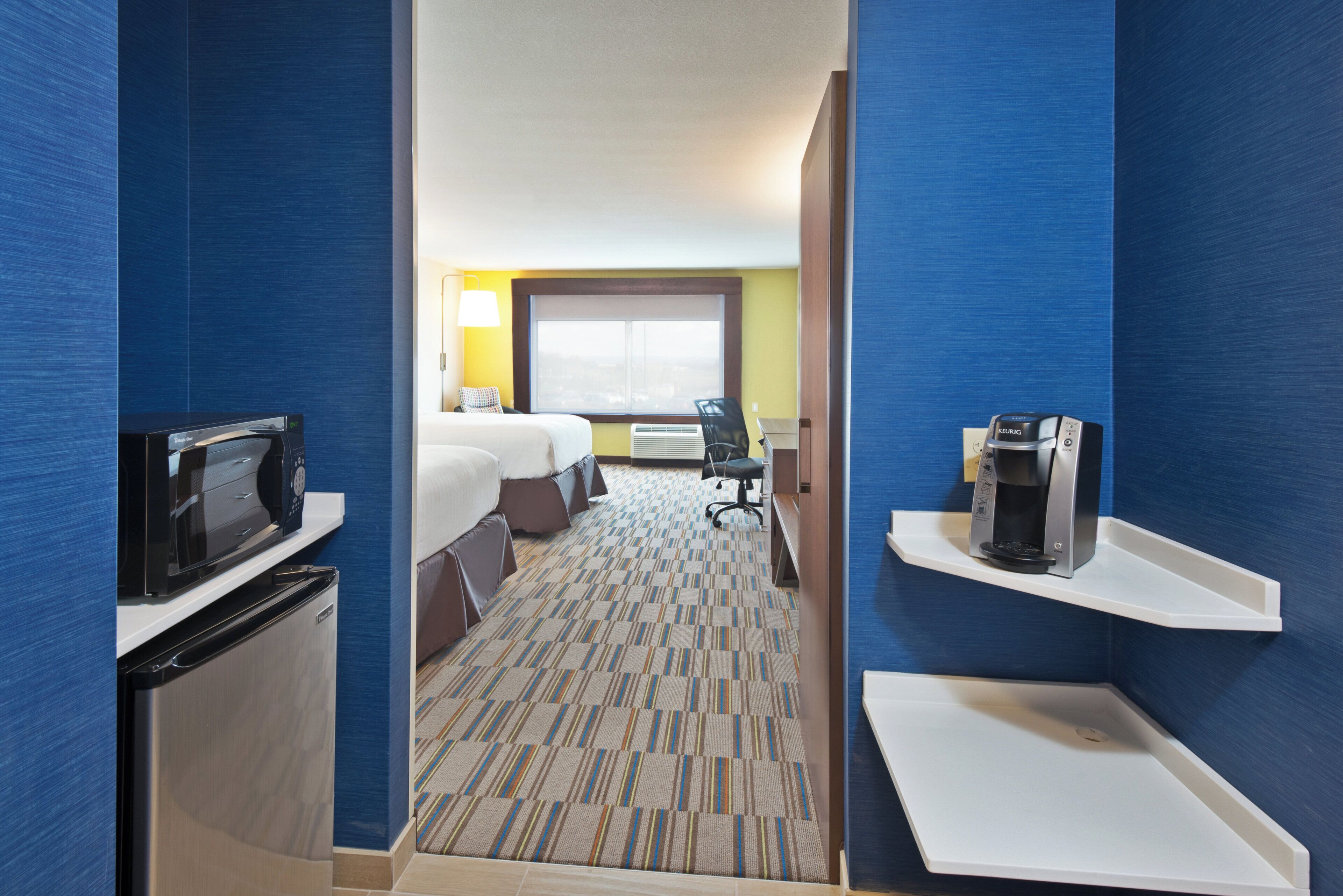A hotel room with a bed, a desk, a chair, a microwave, a coffee maker, and a refrigerator.