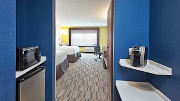 A hotel room with a bed, a desk, a chair, a microwave, a coffee maker, and a refrigerator.
