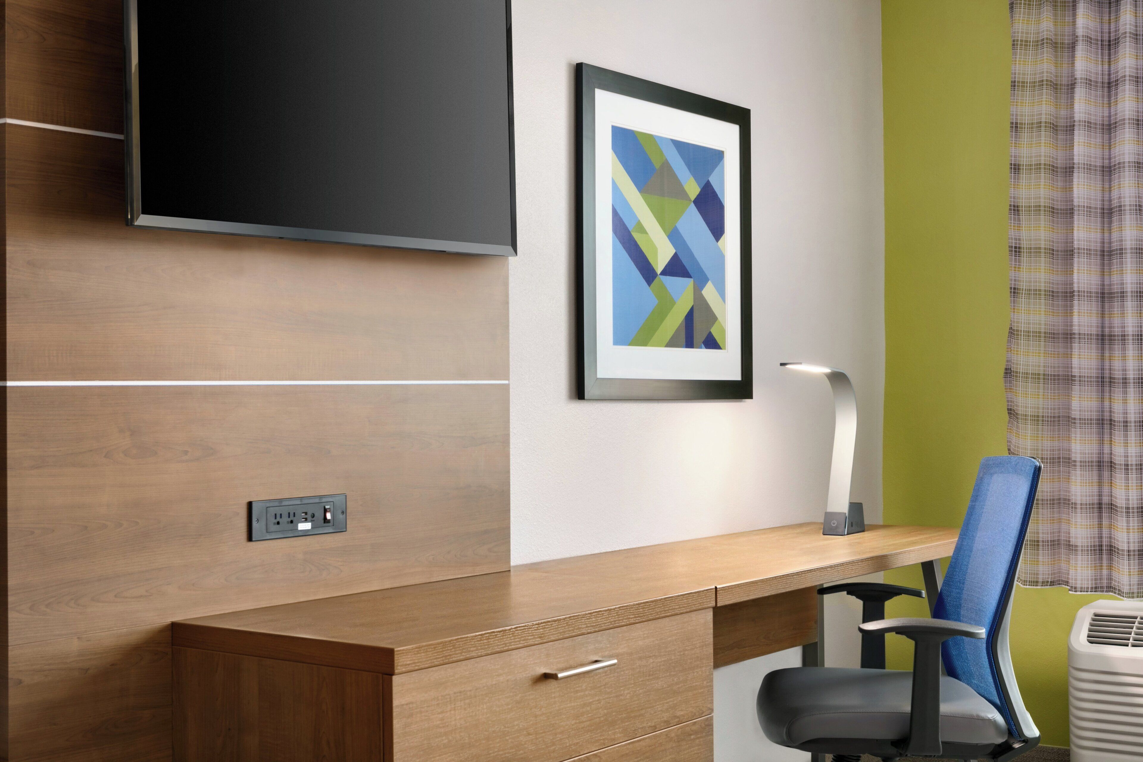A modern office desk with a chair, a mounted flat-screen TV, a framed abstract art piece, and a power outlet.