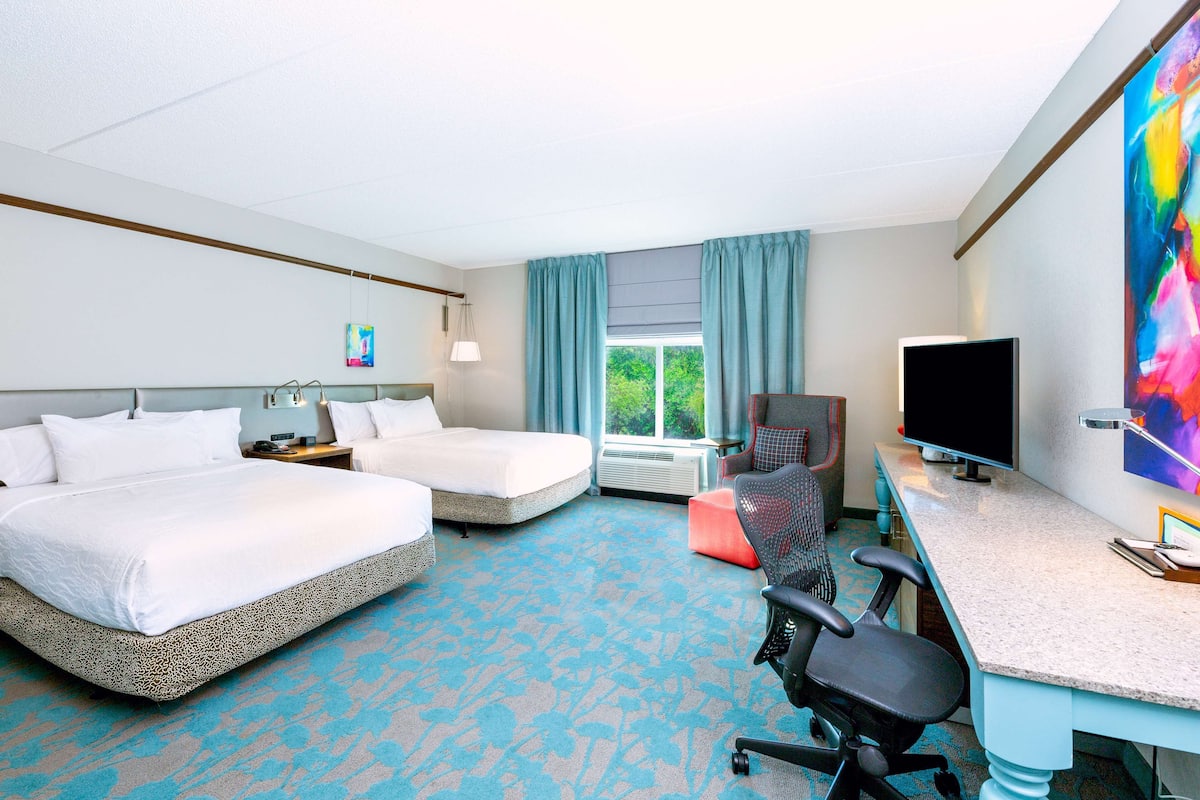 A hotel room with two beds, a desk with a computer, a TV, and a large window with curtains.