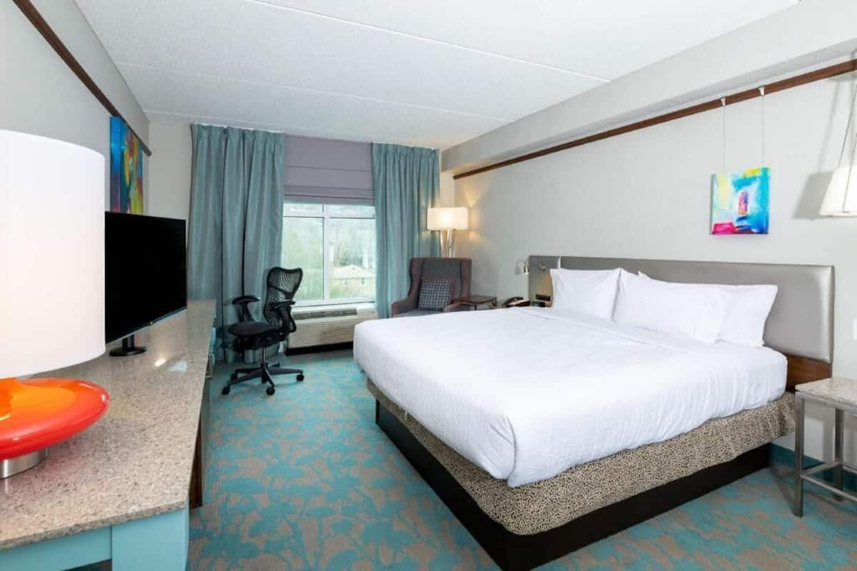 A hotel room with a bed, desk, chair, TV, and artwork.