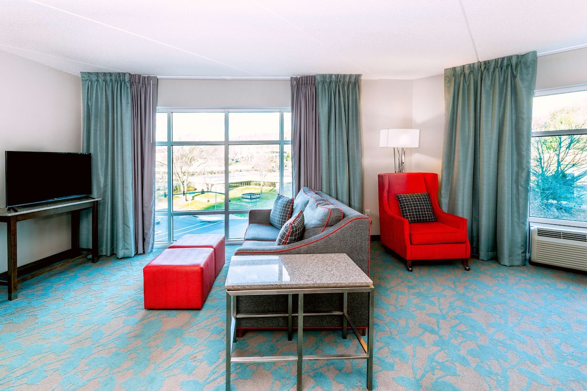 A hotel room with a large window, a sofa, a red armchair, a TV, and a small table.
