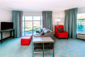 A hotel room with a large window, a sofa, a red armchair, a TV, and a small table.
