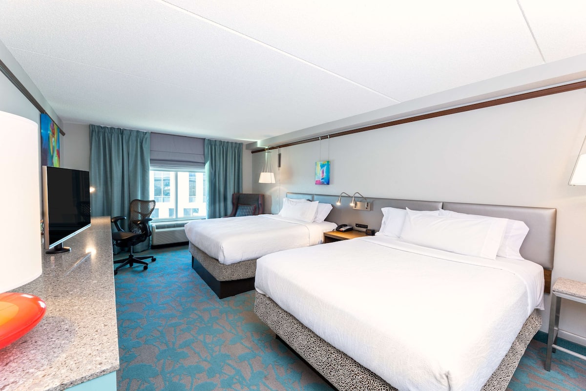 A hotel room with two beds, a desk, a TV, and a window with curtains.