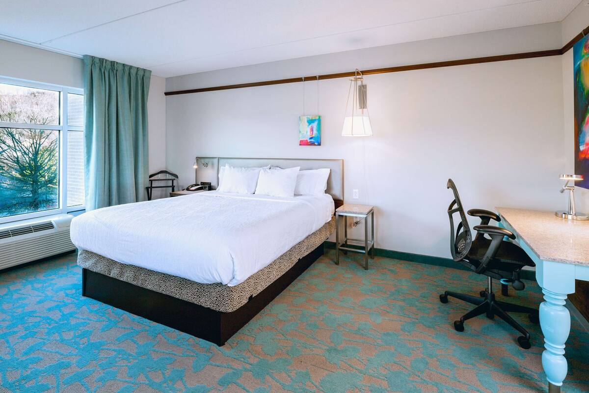 A hotel room with a large bed, a desk, a chair, and a window with curtains.