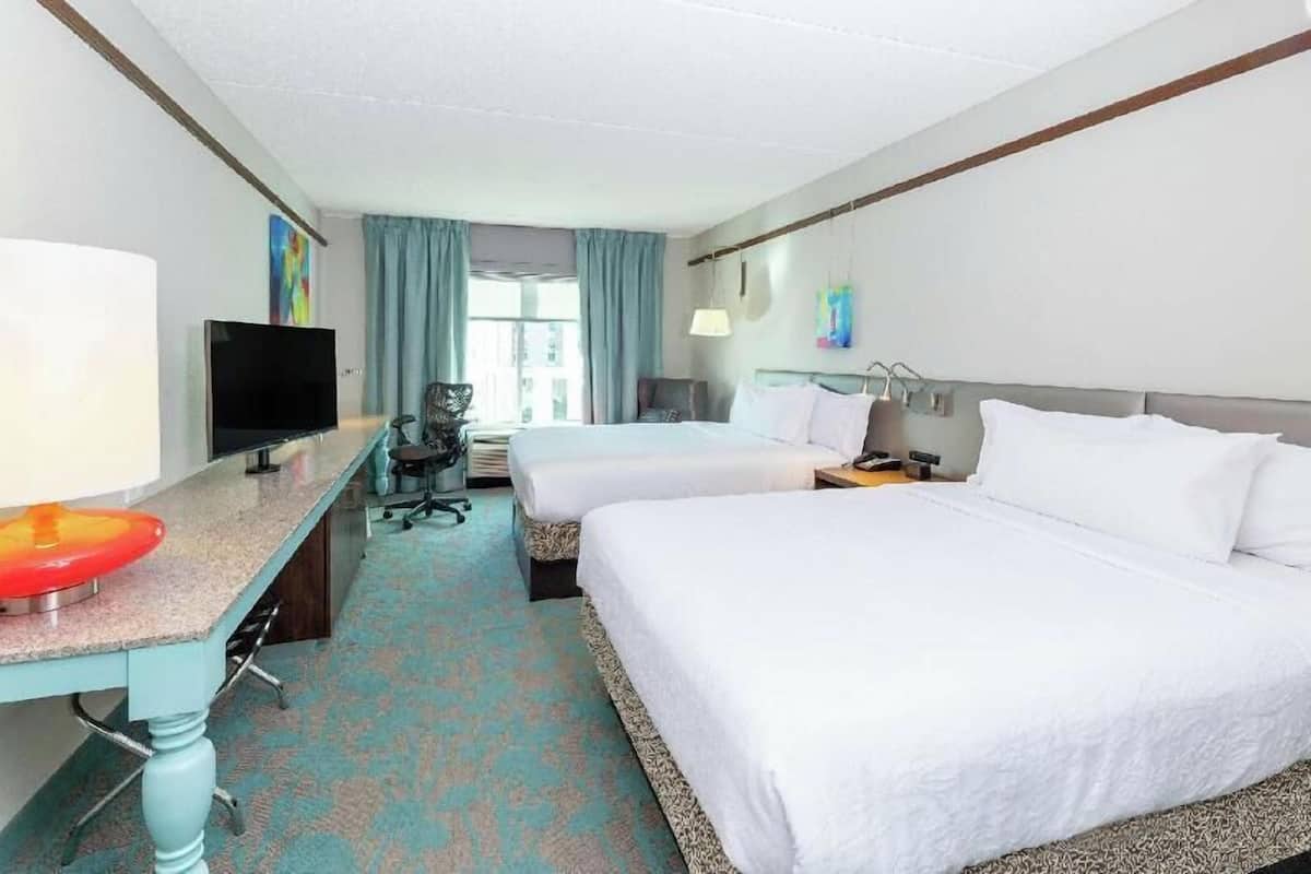 A hotel room with two beds, a desk with a TV, a chair, and a window with curtains.