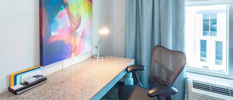 Habitación de hotel con escritorio, silla, equipo de aire acondicionado y un cuadro abstracto grande en la pared.