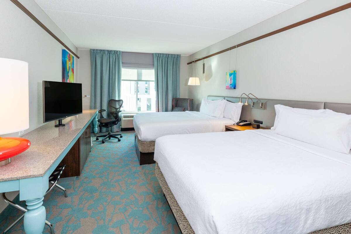 A hotel room with two beds, a desk with a flat-screen TV, a chair, and a window with curtains.