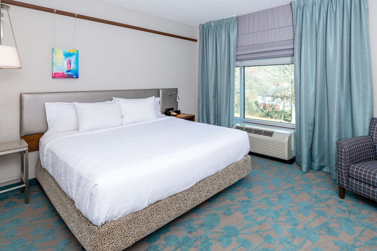 A hotel room with a large bed, a nightstand, a chair, a window with curtains, and a wall-mounted air conditioning unit.