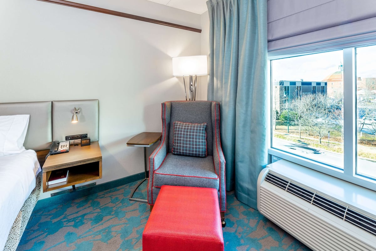 A hotel room with a bed, a desk with a lamp, a chair with a checkered cushion, a red ottoman, and a large window with a view of the outside.