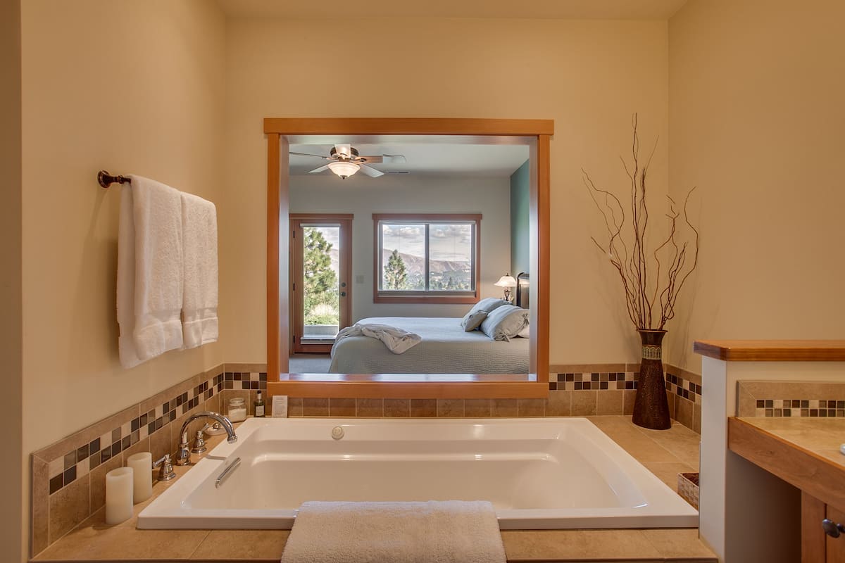A bathroom with a large bathtub, a mirror, and a view into a bedroom with a bed and a window.