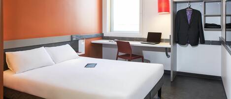 A hotel room with a bed, a desk, a chair, a laptop, a red lamp, and a hanging coat.