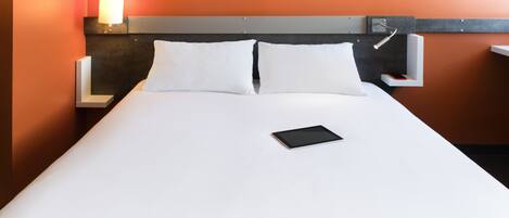 A neatly made bed with white bedding and pillows, a bedside table with a lamp, and a tablet on the bed.