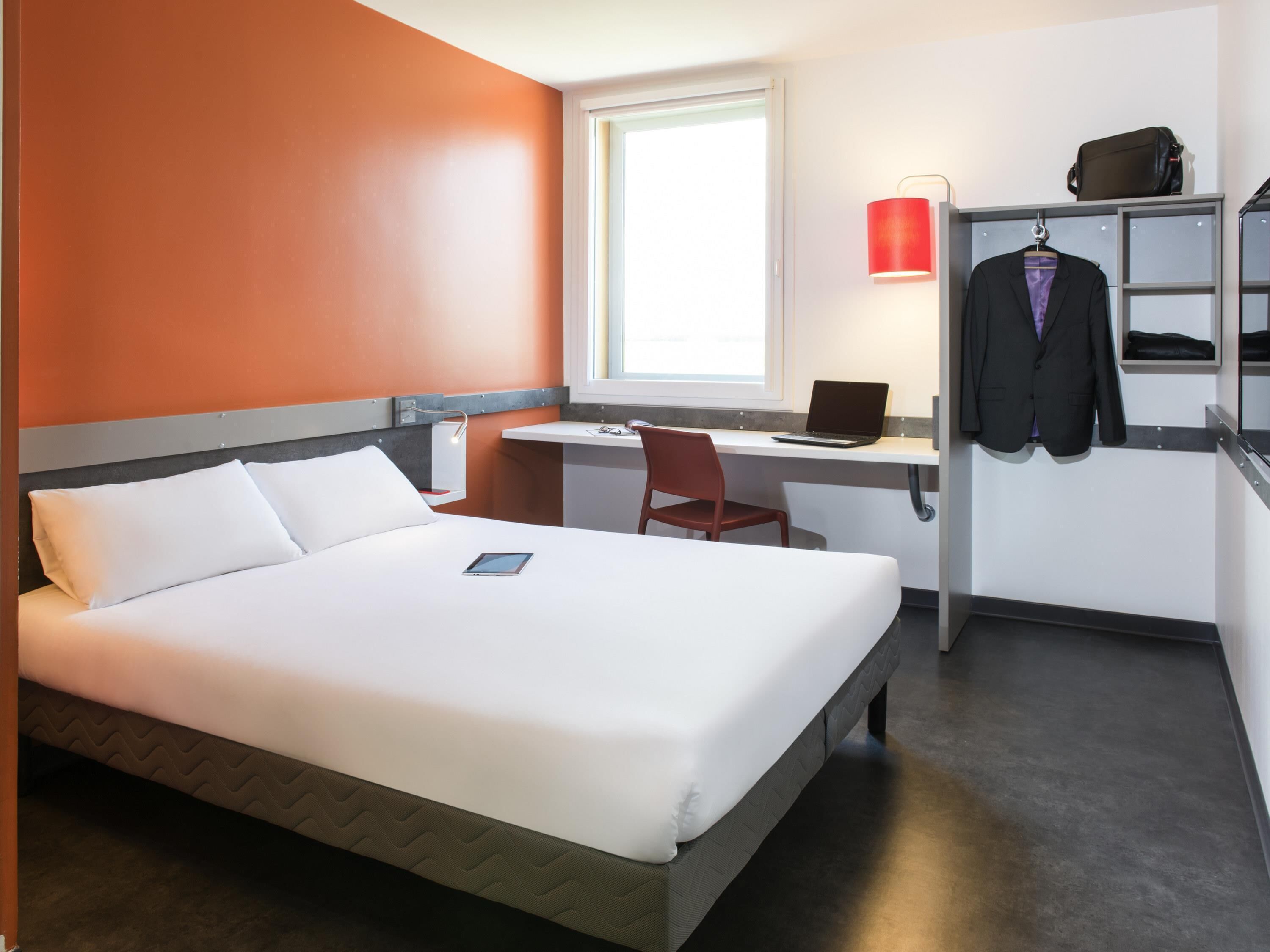 A hotel room with a bed, desk, chair, laptop, and a hanging coat.